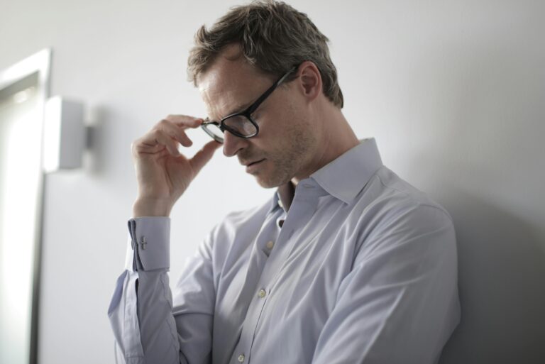 Why do you keep waking up at 3am? An adult man in a shirt and glasses appears stressed while leaning against a wall indoors.