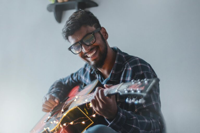 Stress reduction techniques, A smiling man joyfully playing an acoustic guitar with fairy lights indoors.